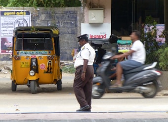 A traffic policeman stands on the side of a road near a parked yellow auto-rickshaw. A person on a scooter is passing by the policeman. The background includes a brick wall with posters, some greenery, and a partially visible commercial establishment.