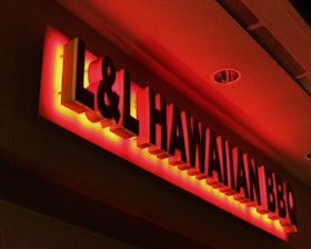 A brightly lit, red neon sign for 'L&L Hawaiian BBQ' is affixed to a wall. The sign is three-dimensional with bold letters and a warm glow emanating from the back of the letters, casting a red hue on the surrounding wall and ceiling.