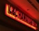 A brightly lit, red neon sign for 'L&L Hawaiian BBQ' is affixed to a wall. The sign is three-dimensional with bold letters and a warm glow emanating from the back of the letters, casting a red hue on the surrounding wall and ceiling.