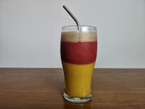 A layered smoothie in a tall glass with three distinct sections of different colors: yellow at the bottom, red in the middle, and a creamy white on top. A metal straw is placed within the drink, and the glass is set on a wooden surface against a plain background.