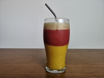 A layered smoothie in a tall glass with three distinct sections of different colors: yellow at the bottom, red in the middle, and a creamy white on top. A metal straw is placed within the drink, and the glass is set on a wooden surface against a plain background.