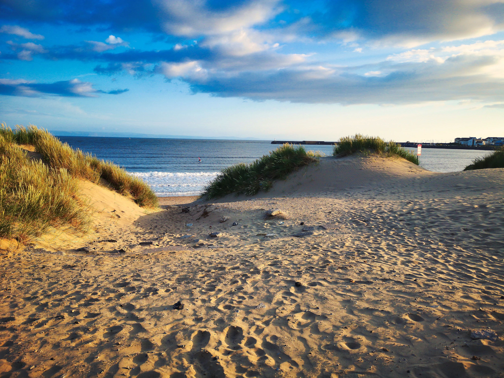 A sandy path winds between grassy dunes toward the ocean under a partly cloudy sky.