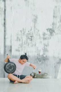 A person with a tied-up hairstyle is sitting cross-legged on the floor. They are holding a large pan in one hand and using a ladle in the other hand, as if pretending to eat from a fountain or decorative water feature that contains some food items. The setting includes a textured wall with a weathered appearance.