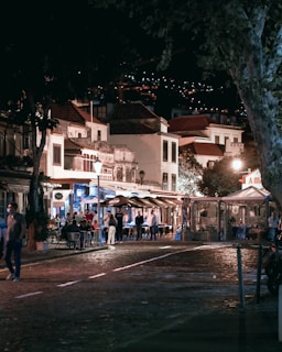 A lively street scene at night with people dining outside at a row of quaint restaurants and cafes. The ambiance is warm with lights illuminated from the buildings and street lamps, creating a cozy atmosphere. The cobblestone street in the foreground adds to the charm of the setting, and trees partially frame the scene.