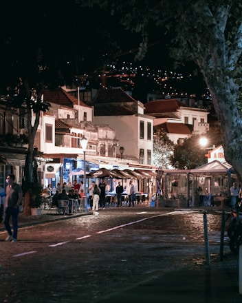 A lively street scene at night with people dining outside at a row of quaint restaurants and cafes. The ambiance is warm with lights illuminated from the buildings and street lamps, creating a cozy atmosphere. The cobblestone street in the foreground adds to the charm of the setting, and trees partially frame the scene.