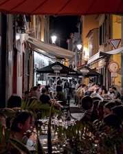 A bustling outdoor dining scene at night with people seated at tables along a narrow street, illuminated by surrounding lights. The atmosphere is lively, and umbrellas with branding provide shelter to some of the tables. The street is flanked by old-style buildings, adding a charming character to the setting.