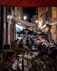 A bustling outdoor dining scene at night with people seated at tables along a narrow street, illuminated by surrounding lights. The atmosphere is lively, and umbrellas with branding provide shelter to some of the tables. The street is flanked by old-style buildings, adding a charming character to the setting.