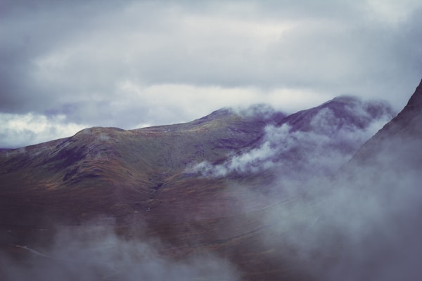 A misty landscape featuring rolling hills partially obscured by low-hanging clouds. The terrain appears rugged and covered in patches of greenery, with a muted color palette that is enhanced by the overcast sky.