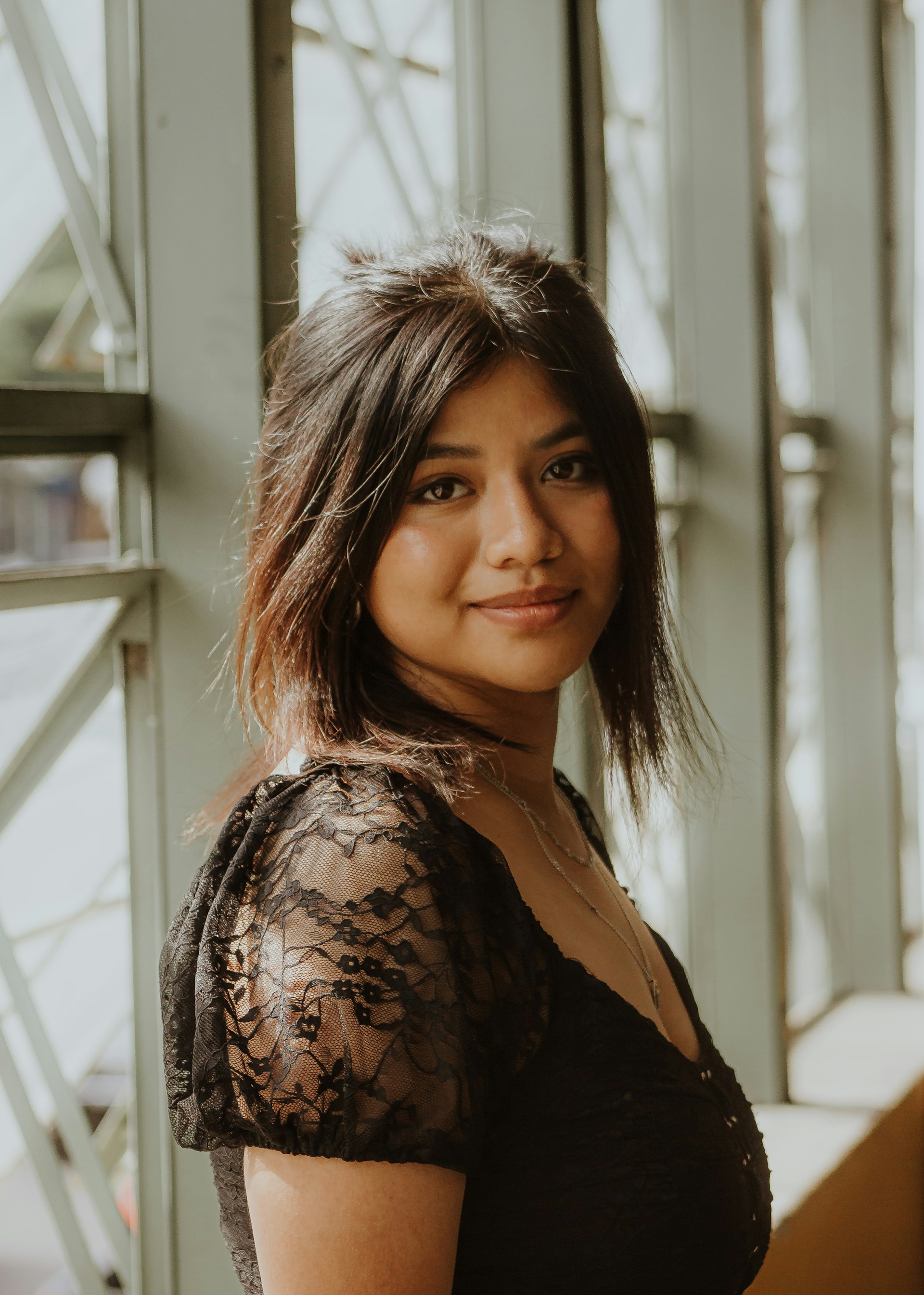 A charming escort standing by a window, natural light highlighting her relaxed and inviting expression.