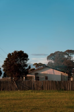 A small house surrounded by trees and a wooden fence under a clear blue sky. The building appears to be a single-story structure, partially illuminated by warm sunlight, creating a peaceful countryside setting.