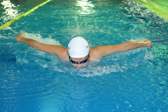 A swimmer performing the butterfly stroke in a swimming pool, wearing a white swim cap and goggles. The water is choppy with splashes created by the swimmer's movement. The lanes in the pool are marked with a rope on the left side.