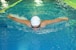 A swimmer performing the butterfly stroke in a swimming pool, wearing a white swim cap and goggles. The water is choppy with splashes created by the swimmer's movement. The lanes in the pool are marked with a rope on the left side.