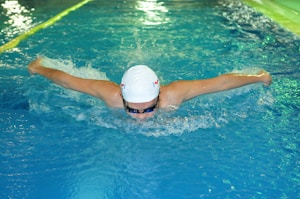A swimmer performing the butterfly stroke in a swimming pool, wearing a white swim cap and goggles. The water is choppy with splashes created by the swimmer's movement. The lanes in the pool are marked with a rope on the left side.