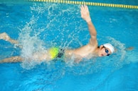 A swimmer is captured in mid-stroke while performing a freestyle swim in a pool. The individual is wearing a white swim cap and blue goggles, with a vibrant green and blue swimsuit. Water splashes energetically around them, indicating motion and effort. A yellow lane divider is visible just above the swimmer.