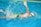 A swimmer is captured in mid-stroke while performing a freestyle swim in a pool. The individual is wearing a white swim cap and blue goggles, with a vibrant green and blue swimsuit. Water splashes energetically around them, indicating motion and effort. A yellow lane divider is visible just above the swimmer.