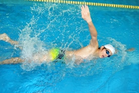 A swimmer is captured in mid-stroke while performing a freestyle swim in a pool. The individual is wearing a white swim cap and blue goggles, with a vibrant green and blue swimsuit. Water splashes energetically around them, indicating motion and effort. A yellow lane divider is visible just above the swimmer.