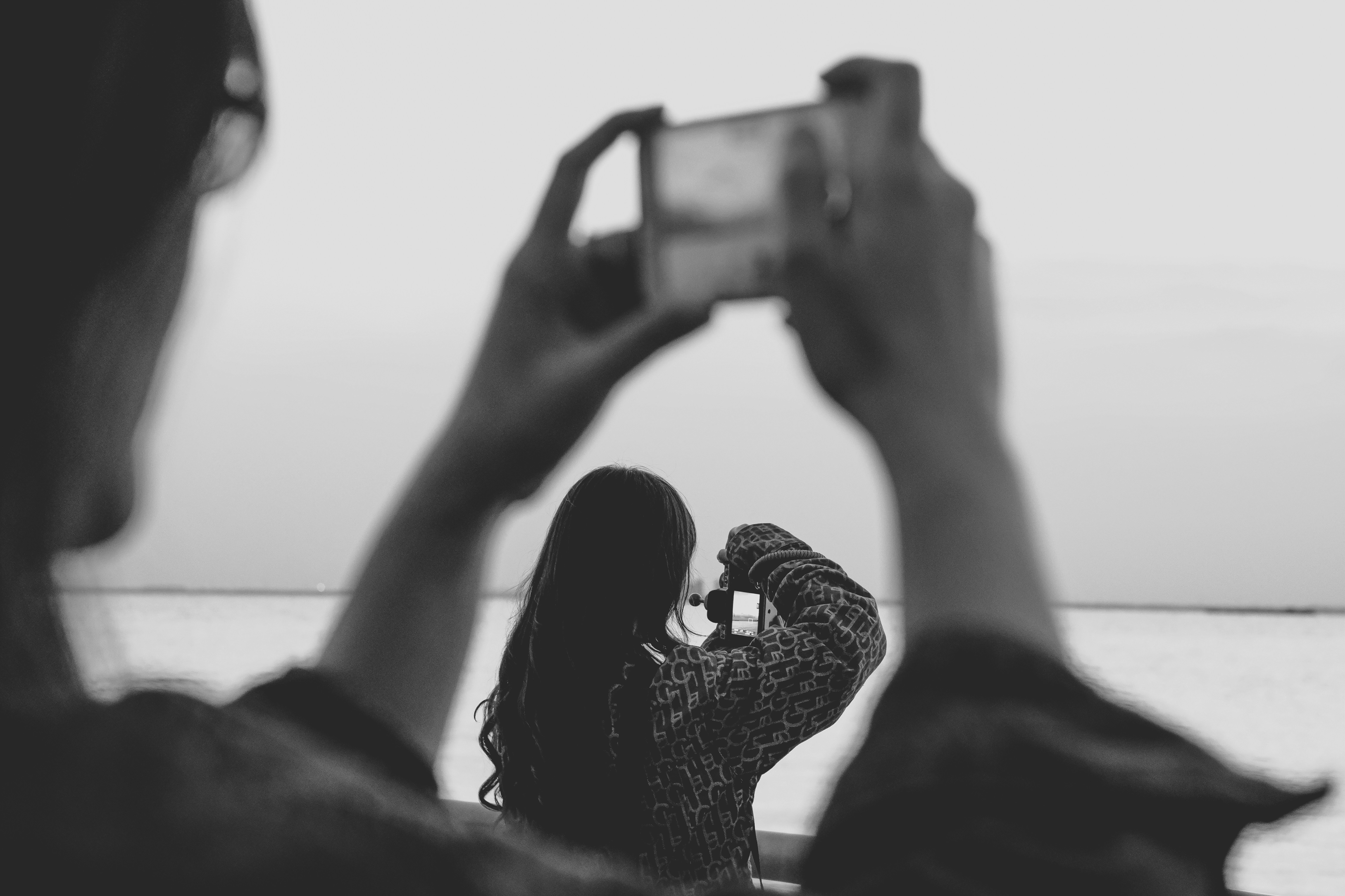 A happy young couple sitting together outdoors, smiling as the man holds up a smartphone to take a candid photo of his girlfriend.