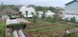 An urban garden with neatly arranged plots of soil, surrounded by various residential and commercial buildings. There are small greenhouses made of transparent plastic and a few trees scattered around the area. The garden is bordered by fences, and in the background, a mix of vegetation and rooftops is visible under a cloudy sky.