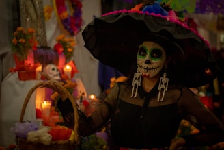 A person is dressed in a traditional Day of the Dead costume, featuring a large black hat adorned with colorful flowers. The face is intricately painted with a skull design, and there is a basket containing vibrant paper flowers. In the background, an altar is decorated with candles, marigold flowers, and a skull.