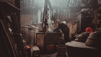 An elderly person is working in a rustic, dimly lit kitchen filled with wooden furnishings and various kitchen tools. The environment appears old-fashioned with wooden walls and a ceiling where light filters through. A large pot is placed at the center where the person is focused, surrounded by baskets and other cooking utensils. Vegetables are visible on the side, and the overall ambiance suggests a traditional setting.