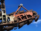 A large rusted industrial machine with intricate gears and mechanisms is set against a clear blue sky. The metal parts are weathered, showing signs of aging and use, with a mix of brown and red oxidation visible. The machine's design suggests it is used for heavy-duty mechanical work or construction.