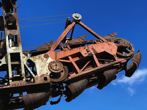 A large rusted industrial machine with intricate gears and mechanisms is set against a clear blue sky. The metal parts are weathered, showing signs of aging and use, with a mix of brown and red oxidation visible. The machine's design suggests it is used for heavy-duty mechanical work or construction.