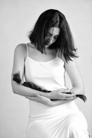 A woman with shoulder-length hair is seated, wearing a sleeveless white dress, and holding a long braid of hair in her arms while looking downward.