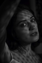 A monochromatic image of a woman gazing directly at the camera. The light creates a pattern of shadows on her face and body, adding to the dramatic effect. She is wearing a sleeveless top and has slightly parted lips with a neutral expression.