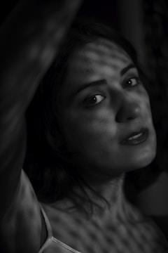 A monochromatic image of a woman gazing directly at the camera. The light creates a pattern of shadows on her face and body, adding to the dramatic effect. She is wearing a sleeveless top and has slightly parted lips with a neutral expression.