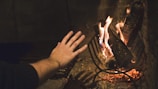 A hand is extended towards a crackling fire inside a fireplace. The firewood is positioned on a metal grate with flames and embers glowing warmly. The background is a dimly lit, rustic setting, creating a cozy atmosphere.