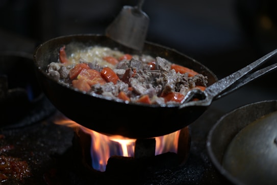 A pan filled with cooking food, including chunks of meat and slices of tomatoes, is positioned over an open flame. The ingredients appear to be sizzling, with visible steam and juices bubbling. The metal spatula resting on the side of the pan suggests cooking in progress.
