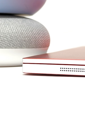A close-up of a smart speaker with a fabric texture sits adjacent to a closed red laptop. The laptop has a sleek design with visible speaker holes on its side.
