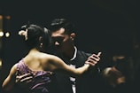 A couple is engaged in a close, intimate dance, captured in low-light conditions. The woman is wearing a purple dress with floral details, while the man is dressed in a dark suit. Their expressions are focused and serene, contributing to an atmosphere of elegance and grace.
