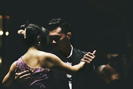 A couple is engaged in a close, intimate dance, captured in low-light conditions. The woman is wearing a purple dress with floral details, while the man is dressed in a dark suit. Their expressions are focused and serene, contributing to an atmosphere of elegance and grace.
