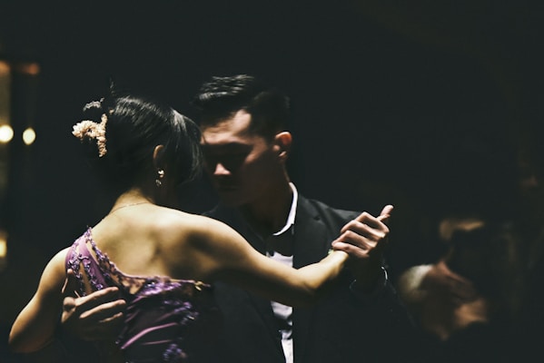 A couple is engaged in a close, intimate dance, captured in low-light conditions. The woman is wearing a purple dress with floral details, while the man is dressed in a dark suit. Their expressions are focused and serene, contributing to an atmosphere of elegance and grace.