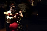 A couple is dancing in a dimly lit setting, with the woman in a red dress and the man in a white shirt. Their close embrace suggests a tango style dance. Several other couples can be seen dancing in the background, with the ambiance suggesting an intimate, serene gathering.