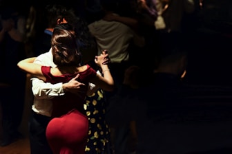 A couple is dancing in a dimly lit setting, with the woman in a red dress and the man in a white shirt. Their close embrace suggests a tango style dance. Several other couples can be seen dancing in the background, with the ambiance suggesting an intimate, serene gathering.