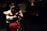 A couple is dancing in a dimly lit setting, with the woman in a red dress and the man in a white shirt. Their close embrace suggests a tango style dance. Several other couples can be seen dancing in the background, with the ambiance suggesting an intimate, serene gathering.