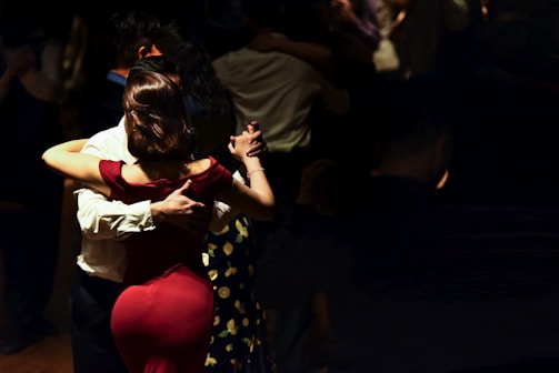 A couple is dancing in a dimly lit setting, with the woman in a red dress and the man in a white shirt. Their close embrace suggests a tango style dance. Several other couples can be seen dancing in the background, with the ambiance suggesting an intimate, serene gathering.