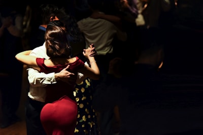 A couple is dancing in a dimly lit setting, with the woman in a red dress and the man in a white shirt. Their close embrace suggests a tango style dance. Several other couples can be seen dancing in the background, with the ambiance suggesting an intimate, serene gathering.