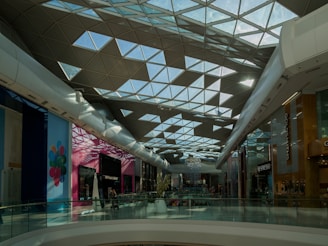 A spacious, modern shopping mall with a geometric glass ceiling allowing natural light to diffuse through. The walls are lined with various stores, including a brightly colored storefront featuring balloons. The floor is polished, and there are people walking or shopping.