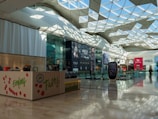 A spacious and well-lit modern shopping mall with a geometrically designed glass ceiling that lets in natural light. Various storefronts are visible, including high-end fashion brands with large advertisements. The floor is polished, reflecting the ambient light, and an escalator and digital signage stand prominently.