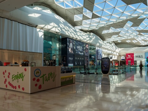 A spacious and well-lit modern shopping mall with a geometrically designed glass ceiling that lets in natural light. Various storefronts are visible, including high-end fashion brands with large advertisements. The floor is polished, reflecting the ambient light, and an escalator and digital signage stand prominently.