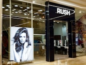 The entrance of a modern hair salon named Rush features sleek glass doors. Inside, the salon is stylishly presented with black chairs and lit mirrors. A large monochrome advertisement poster displays a model with voluminous hair, enhancing the salon's chic interior.