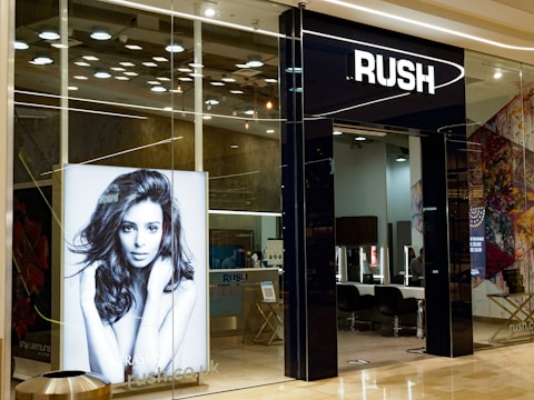 The entrance of a modern hair salon named Rush features sleek glass doors. Inside, the salon is stylishly presented with black chairs and lit mirrors. A large monochrome advertisement poster displays a model with voluminous hair, enhancing the salon's chic interior.