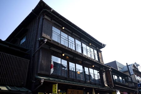 A traditional multi-story Japanese building with wooden architecture and large windows. A Japanese flag hangs on the outside, indicating its cultural significance. The building features intricate wooden details and a classic design typical of historical structures in Japan. The sky is clear and bright in the background.