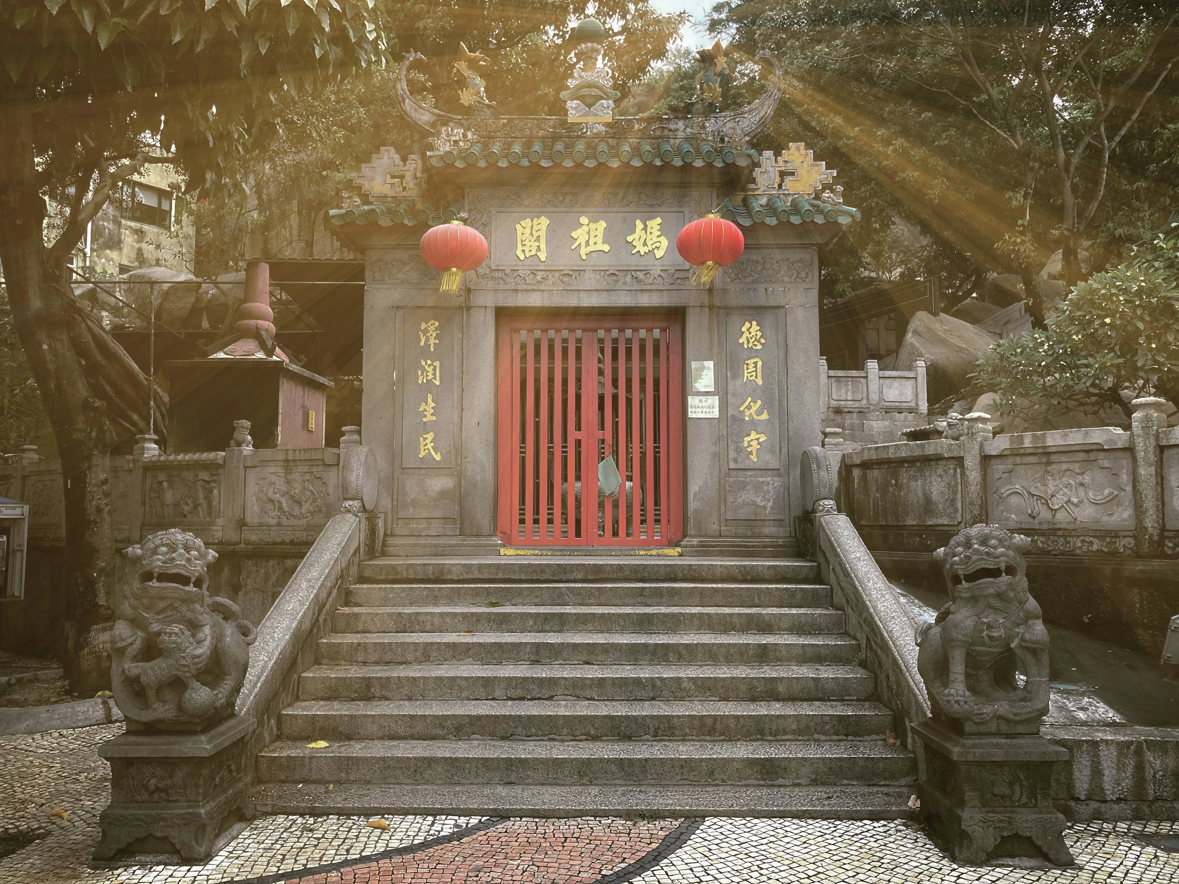 The stone gateway of the A-Ma Temple in Macau, its gilded 媽祖閣 plaque flanked by two red lanterns above a red-slatted door, carved lions on the balustrades and Portuguese wave-patterned paving at the foot of the steps.