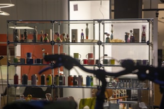 A display shelf with a variety of colorful mugs and figurines is set against a well-lit background in a store. The structure has multiple levels, each with different shaped mugs and artistic figures resembling human forms. A large dark shape in the foreground partially obscures the view.