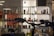 A display shelf with a variety of colorful mugs and figurines is set against a well-lit background in a store. The structure has multiple levels, each with different shaped mugs and artistic figures resembling human forms. A large dark shape in the foreground partially obscures the view.