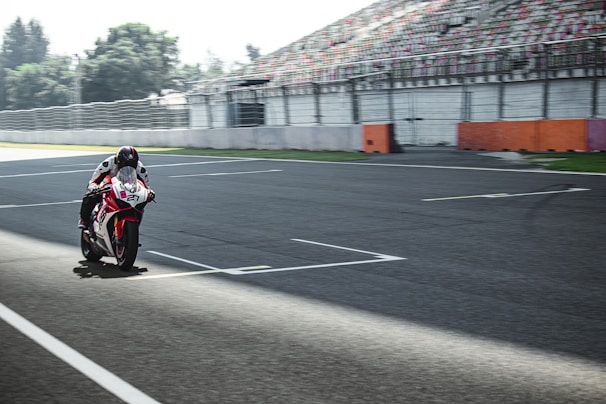A motorcyclist is racing on an empty race track. The rider is leaning forward, wearing full gear, and appears to be speeding on a high-performance sportbike. The track is wide with a backdrop of grandstands and a tree line in the distance.
